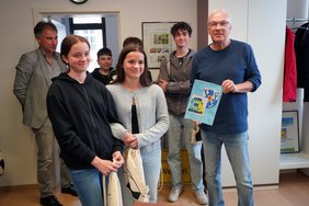 Landrat Udo Recktenwald hält eine Zeichnung, die Schülerinnen ihm gezeichnet haben. Neben und hinter dem Landrat stehen Schüler:innen.