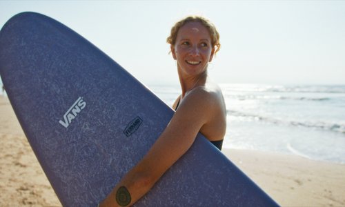 Frau mit Surfbrett unter dem Arm an einem Strand.
